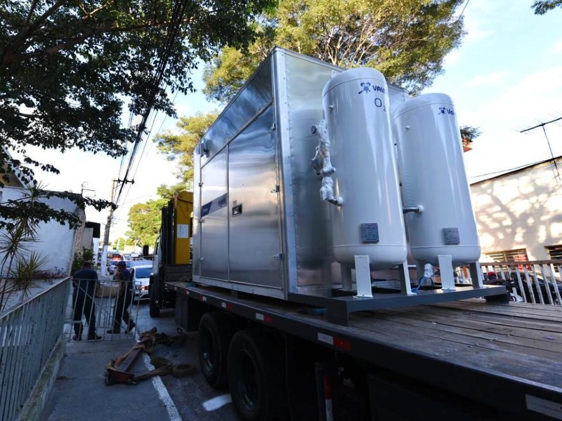 Foto da miniusina de oxigênio do Hospital Dia Tito Lopes. O equipamento contém dois cilindros grandes e um container metálico e está em cima da carroceria de um caminhão, estacionado numa rua arborizada, do lado de fora do hospital